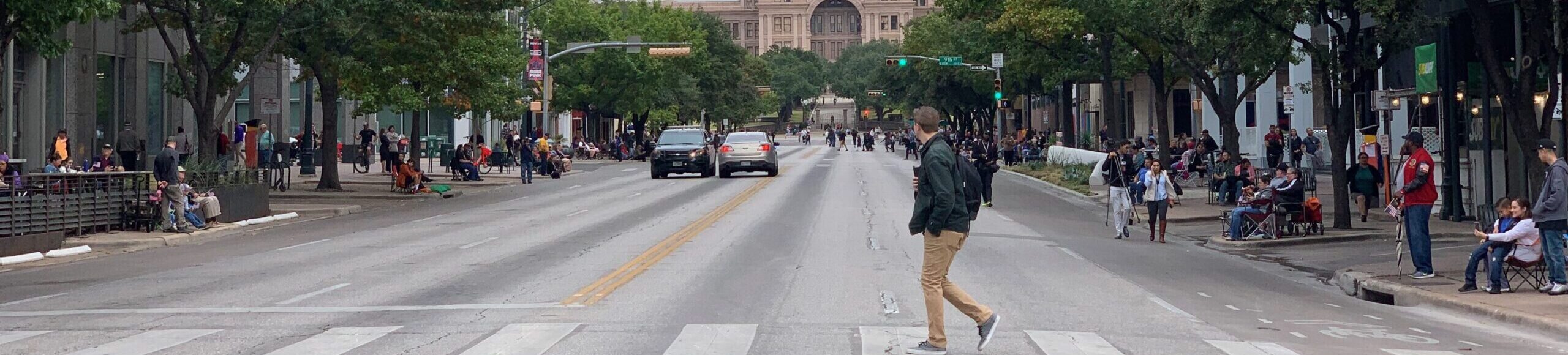 Picture of a guy walking across a crosswalk looking away from the camera at the Texas State Capitol in the background at the end of Congress Avenue.
